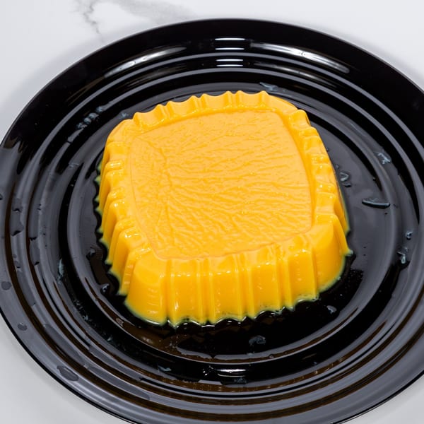 A vibrant yellow mango pudding, molded and glistening, rests on a black ridged plate.